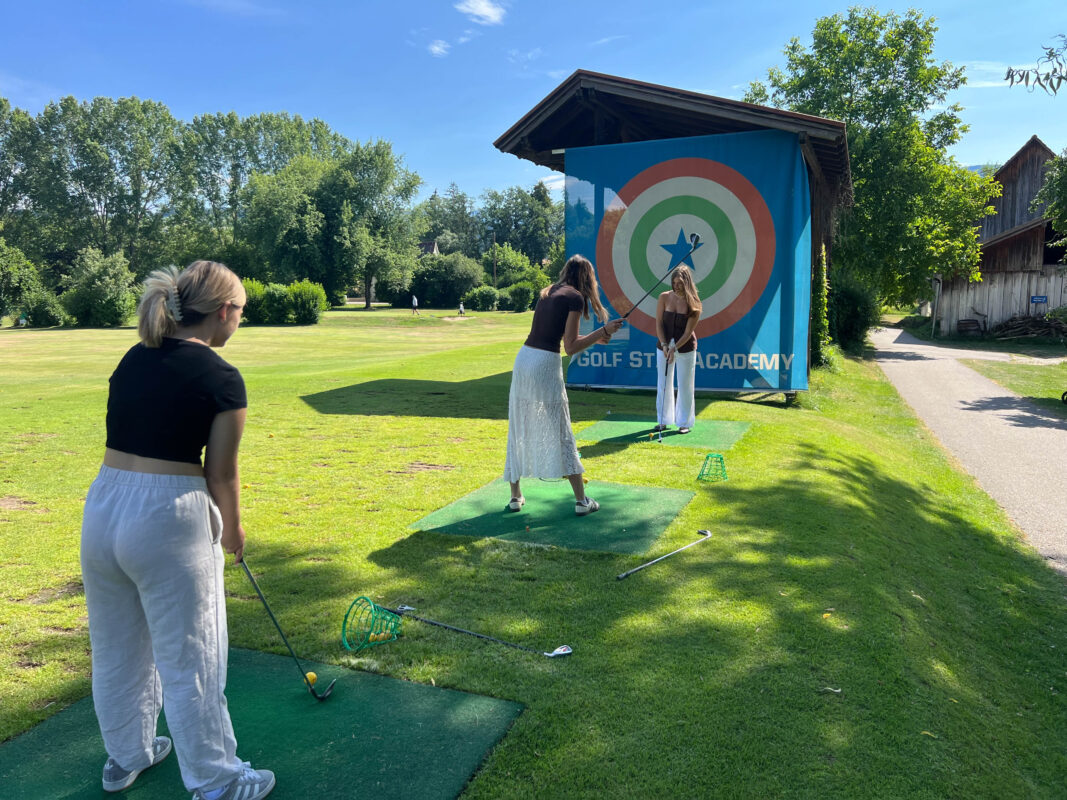 Drei junge Frauen üben Golf auf einer Driving Range vor einer Zielscheibe der Golf Star Academy.